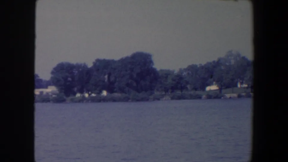 1959 Iowa home-movie frame showing still lake water and a wooded shoreline with scattered small buildings.