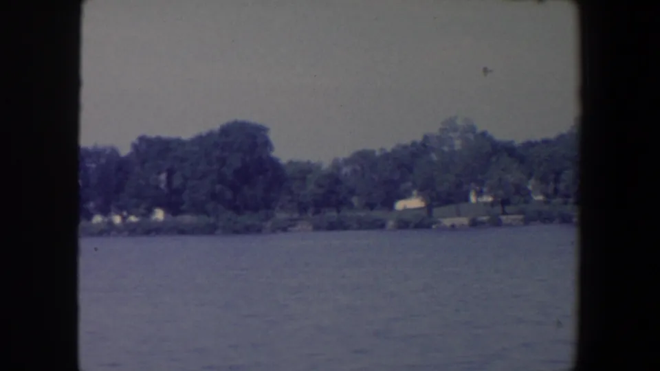 1959 Iowa 8mm frame looking across calm water toward trees and distant lakeside buildings.