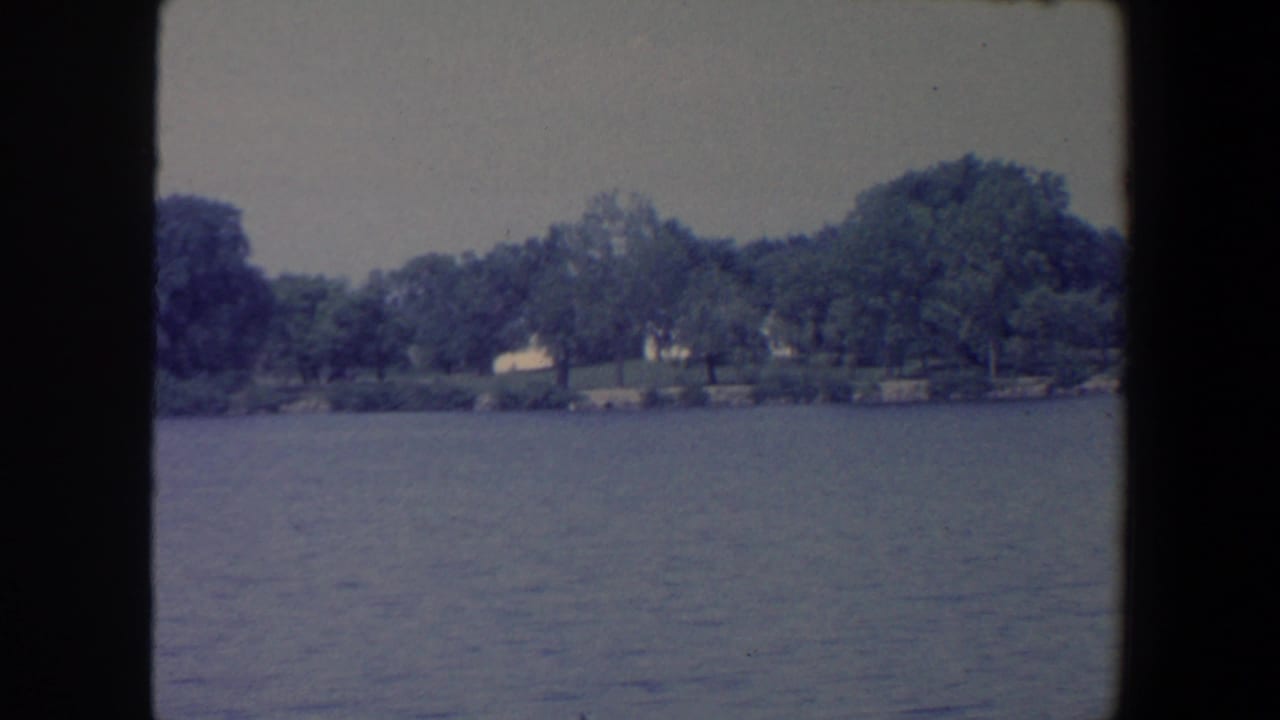 1959 Iowa lake view with rippled water, a wooded shoreline, and pale distant structures.