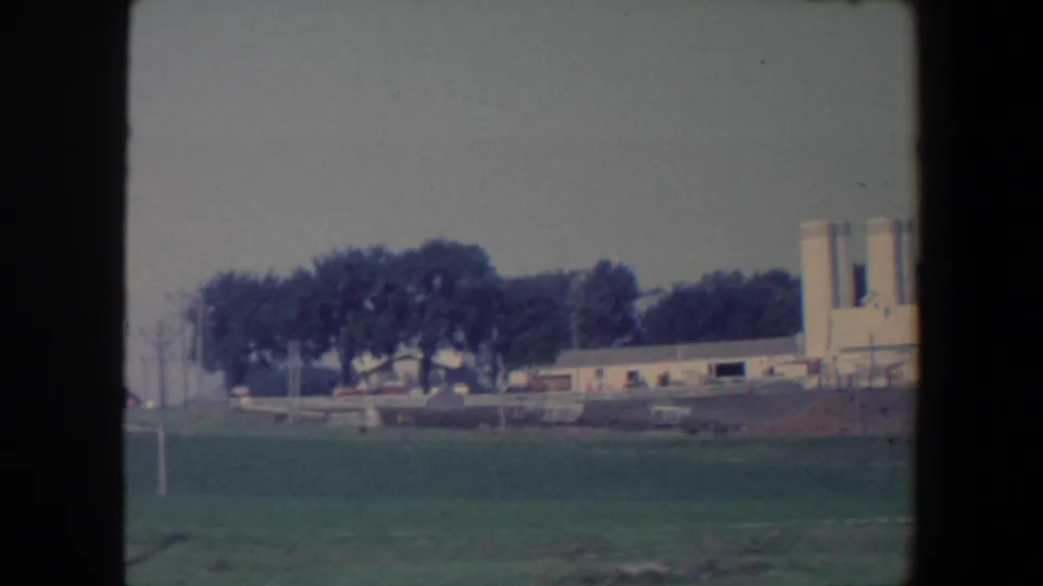 1959 Iowa rural frame showing farm buildings and tall white silos beyond an open green field.