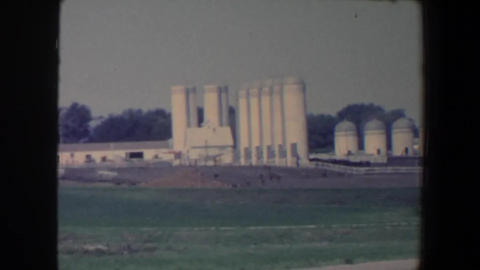 1959 Iowa roadside view of a large agricultural complex with tall white silos, domed tanks, low buildings, fields, and a tree line.