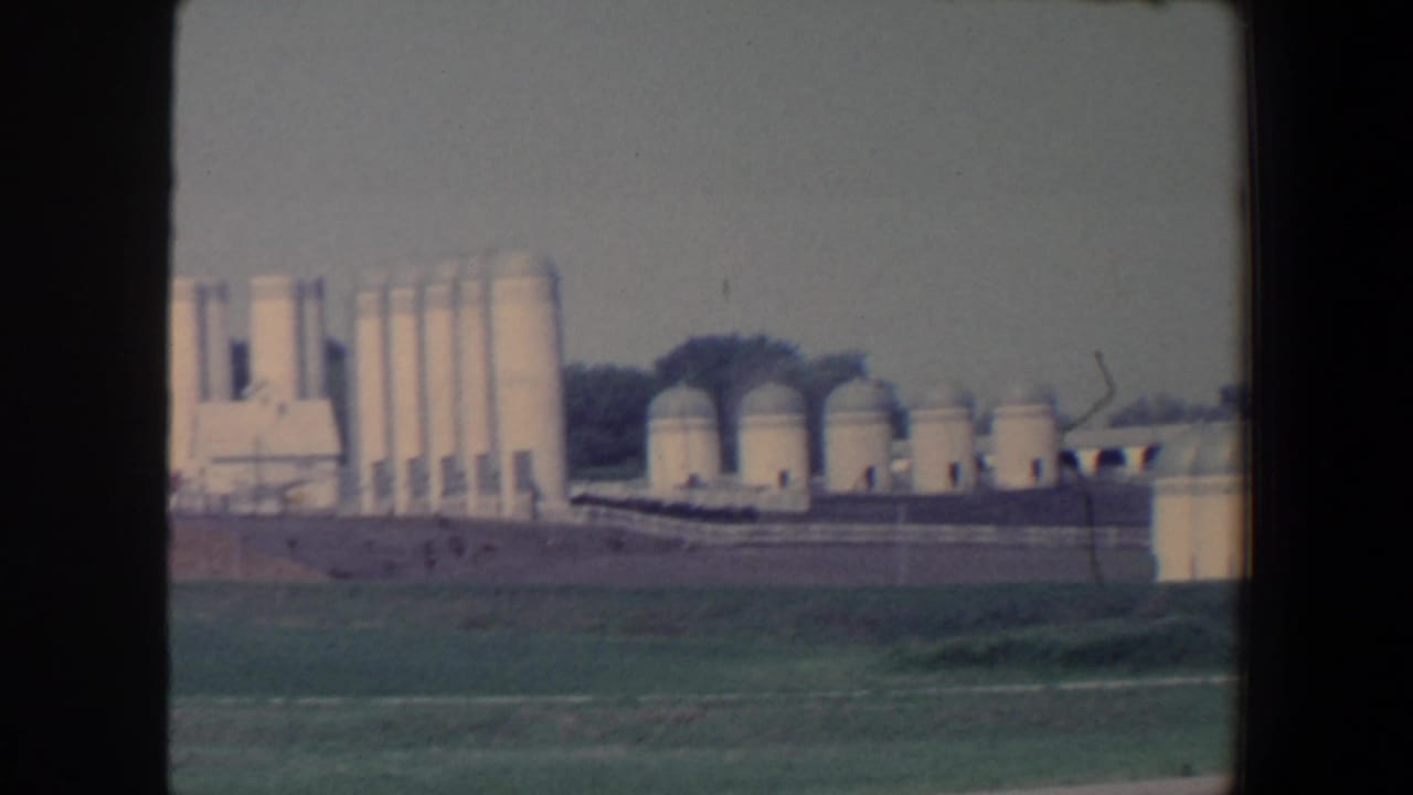 Roadside 1959 Iowa frame showing clustered silos, domed storage tanks, low agricultural structures, green foreground, and distant trees.