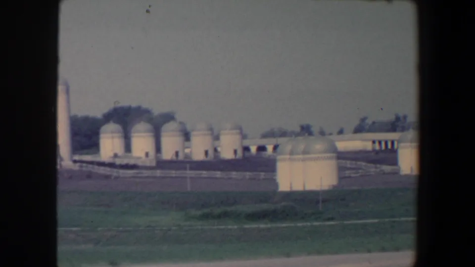 1959 Iowa agricultural complex partly cropped by the moving camera, with domed tanks, low buildings, fields, and a roadside strip visible.