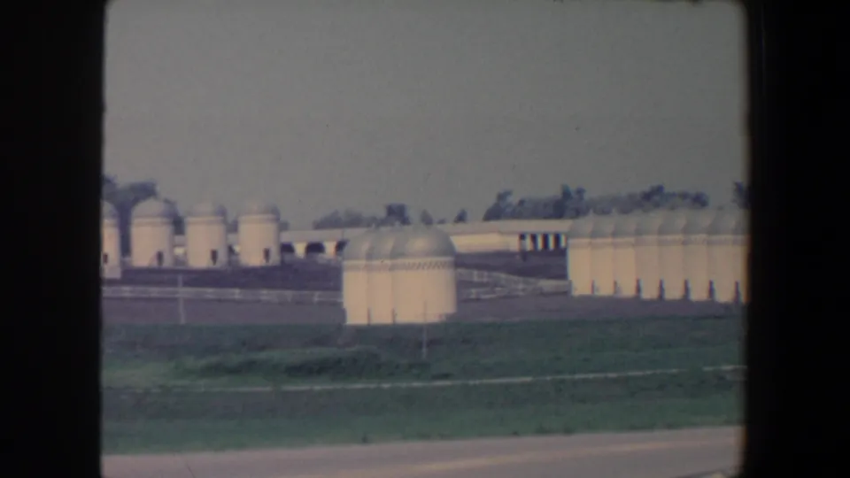 Late frame from the 1959 Iowa clip showing domed agricultural storage tanks, long low buildings, open fields, road edge, and distant trees.