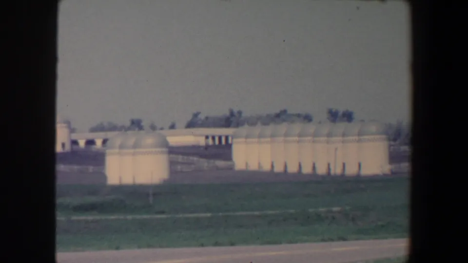 1959 Iowa home-movie frame showing pale silos, domed storage buildings, grass, fencing, and distant trees from a moving roadside view.