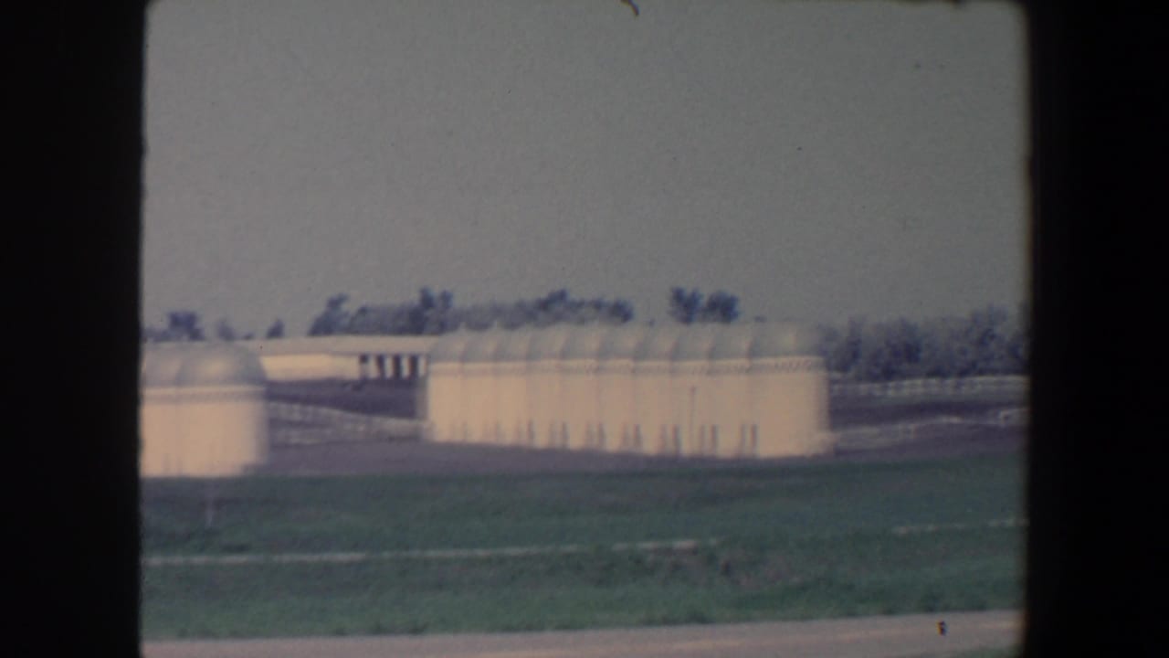 1959 Iowa frame with a row of pale farm silos, white fence lines, open grass, and trees under a hazy sky.