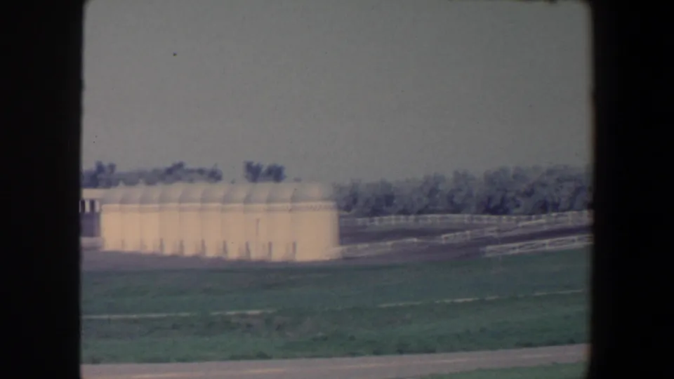 1959 Iowa roadside frame showing cropped white silos, fence rails, pasture-like grass, and a distant tree line.