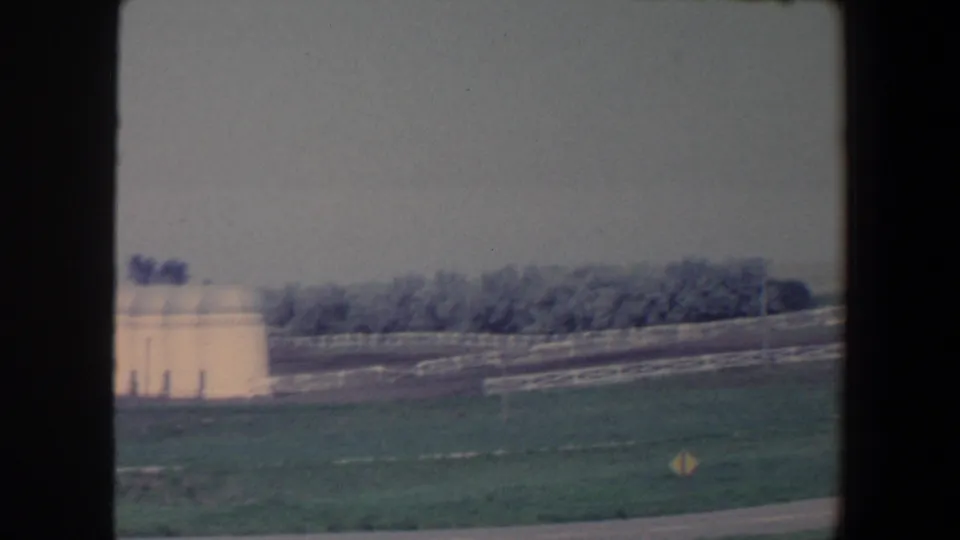 1959 Iowa home-movie frame with white farm structures, long fence lines, open grass, and trees in the background.
