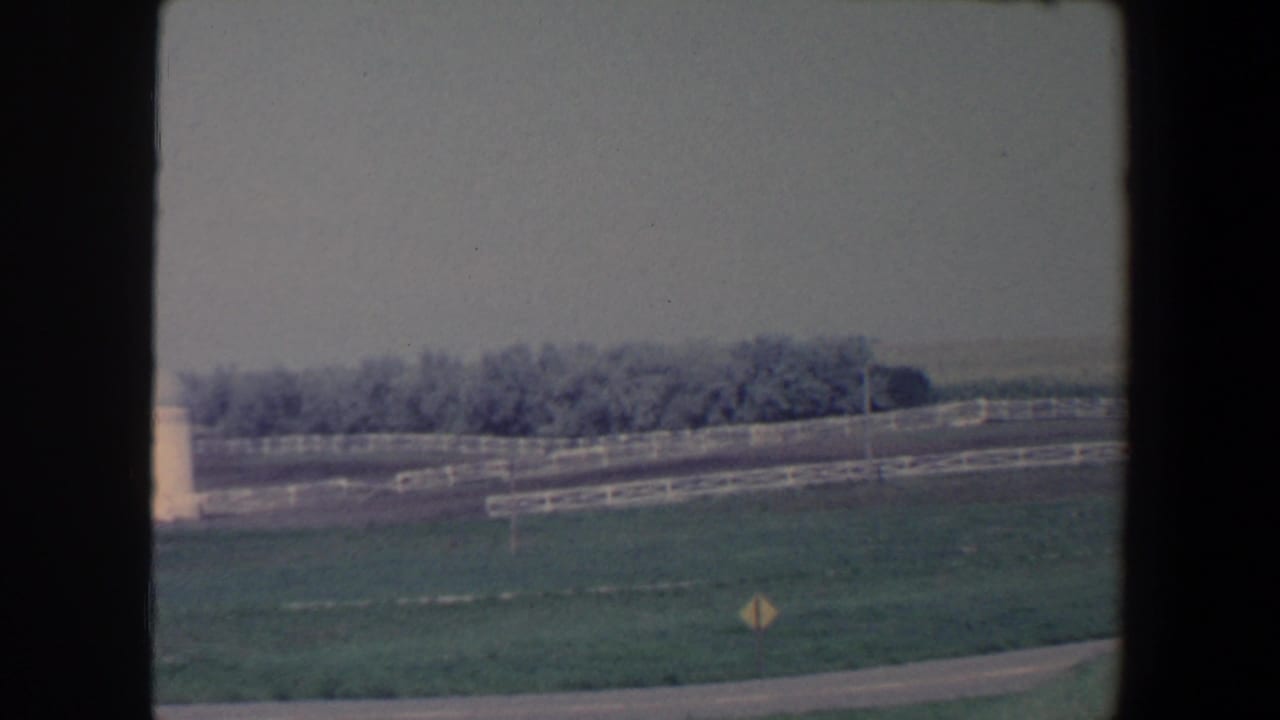 Rural pasture with white fence, roadside curve, yellow sign, and distant treeline.