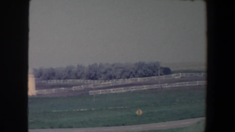 1959 Iowa 8mm frame showing green pasture, white rail fencing, a curving road, yellow roadside sign, and distant trees under a hazy sky.