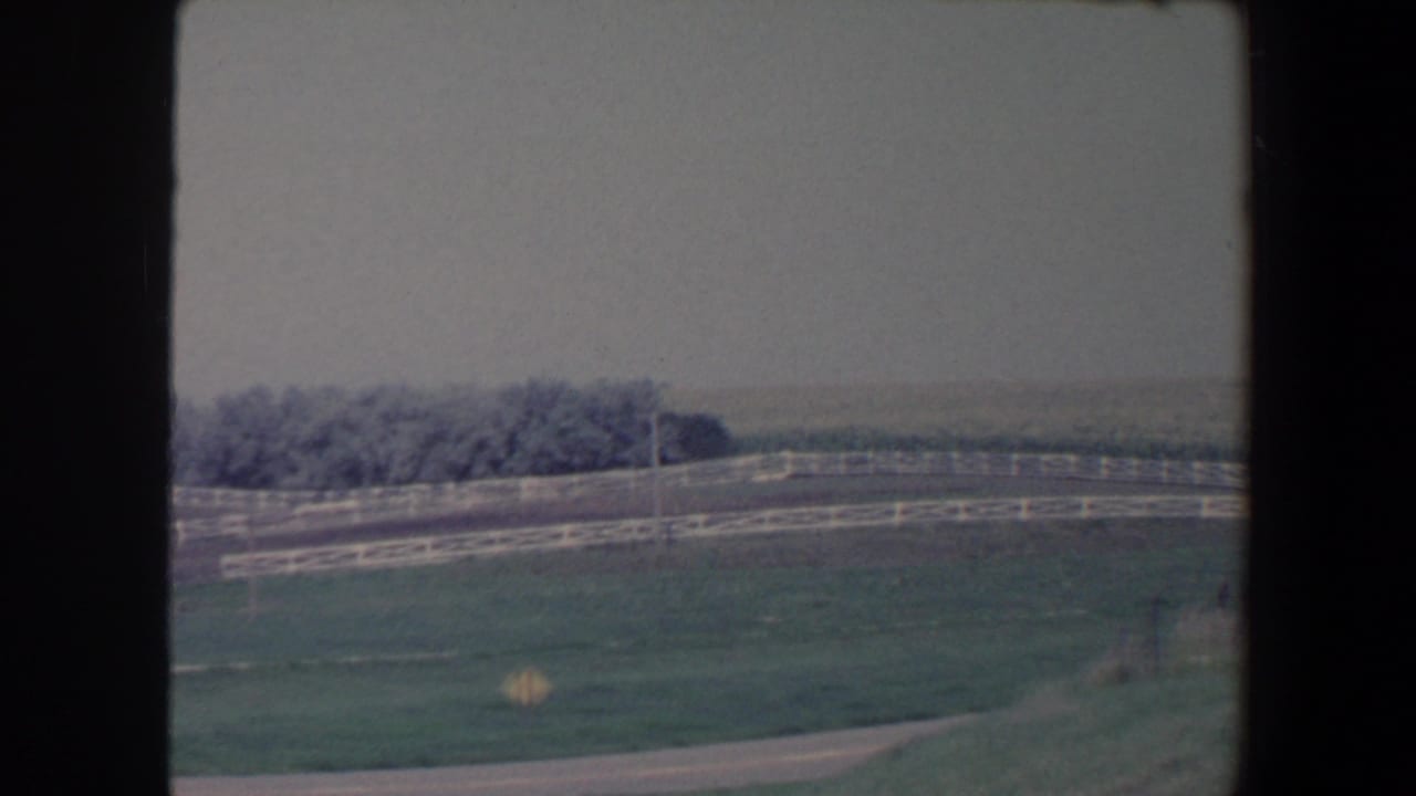 1959 Iowa 8mm frame with a broad pasture, white fencing, a road edge, distant trees, and soft haze across the horizon.