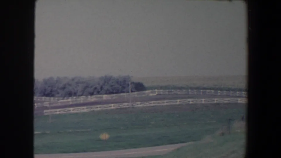 1959 Iowa 8mm frame with a broad pasture, white fencing, a road edge, distant trees, and soft haze across the horizon.