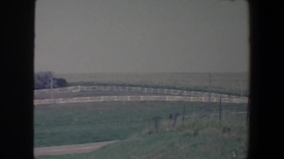 1959 Iowa 8mm frame showing sloped grass, white rail fencing, a road, low brush, and distant rural fields beneath a pale sky.