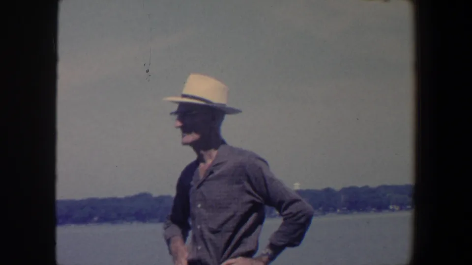 1959 Iowa 8mm frame showing an adult man in a light fedora and dark shirt standing by water, with trees along the far shore.