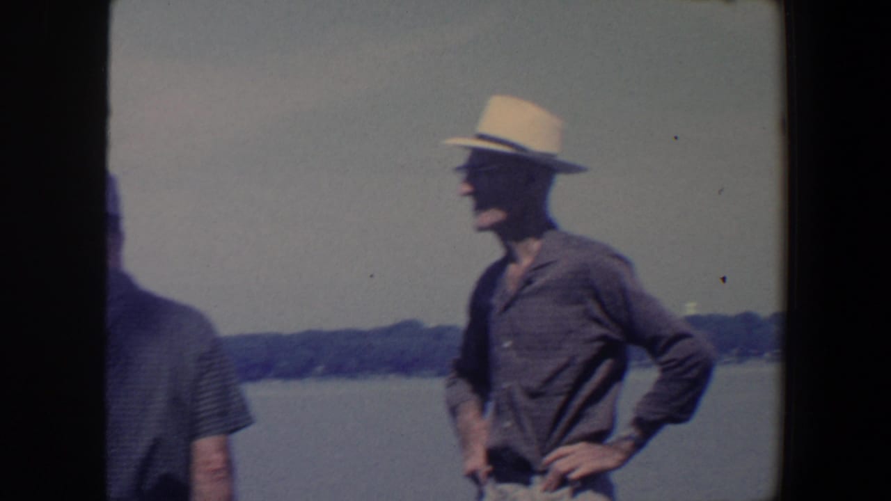 Older man in a light fedora and glasses stands by a lake with another figure partly visible.