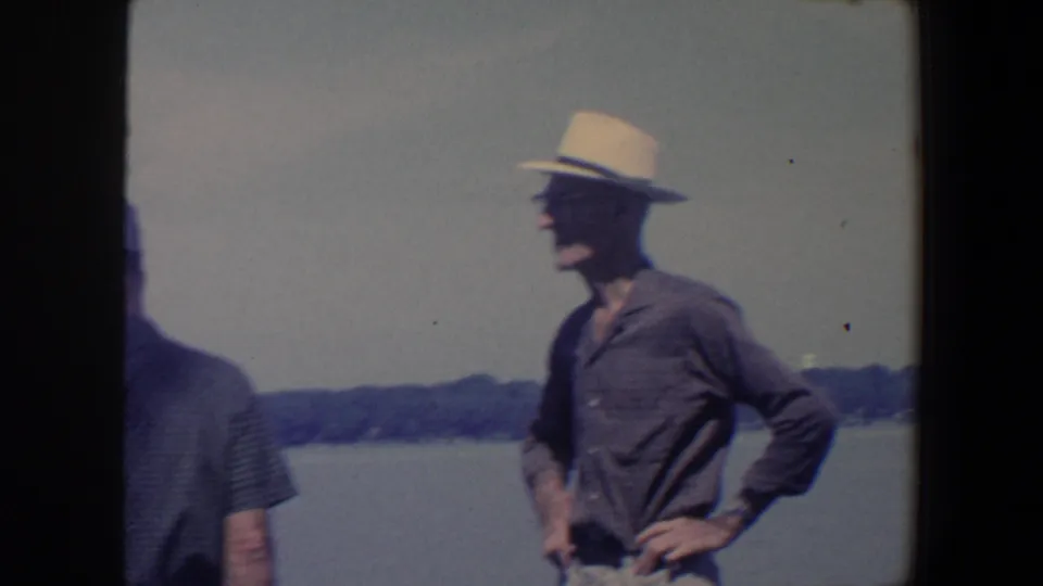 In Iowa 1959 home-movie footage, an older man in a light fedora and glasses stands near lake water with a distant tree-lined shore.