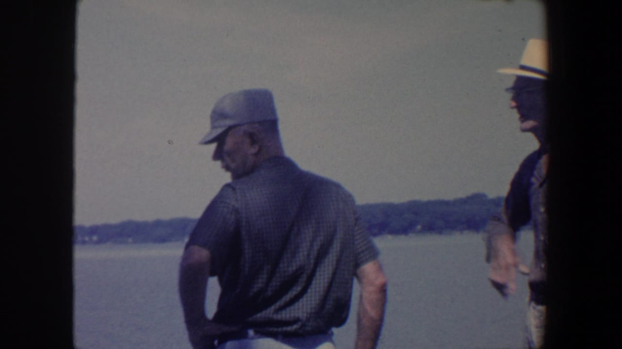 An older man in a dark cap faces the lake while a second older man in a fedora stands partly visible at the right edge.