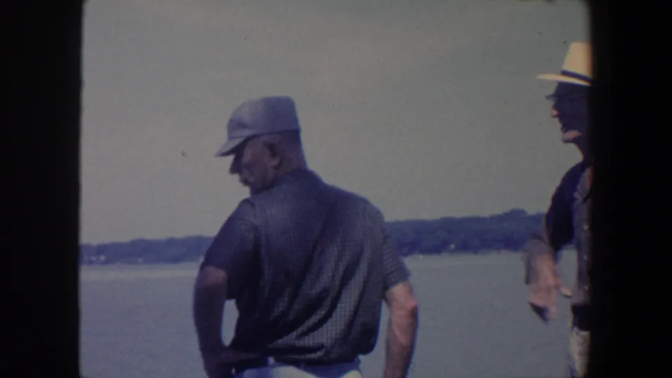 An older man in a dark cap faces the lake while a second older man in a fedora stands partly visible at the right edge.