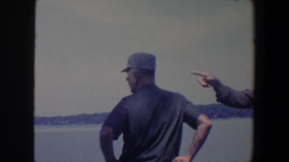 The capped man stands with one hand on his hip as a partly visible arm points toward the distant lakeshore.