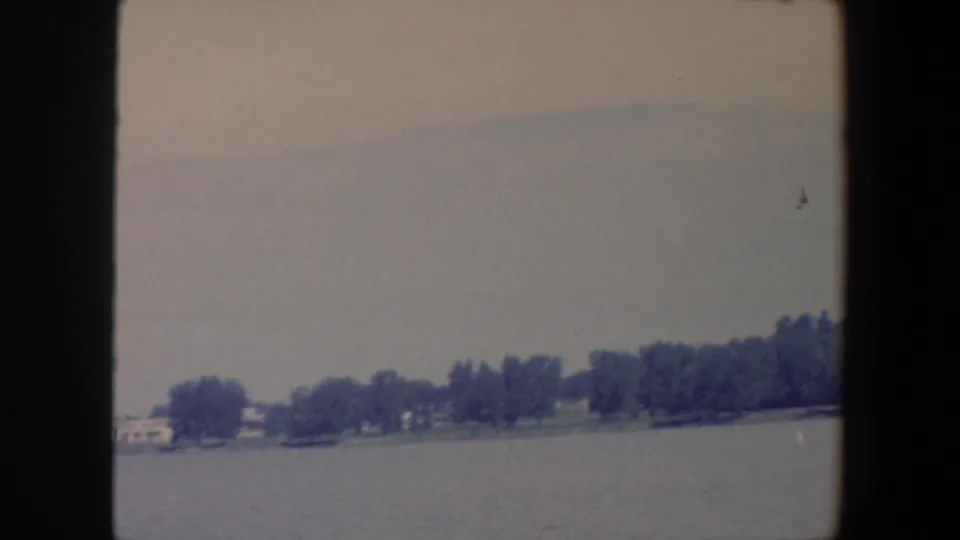 The camera looks across an Iowa lake toward a low tree-lined shore, with a small structure or boat faintly visible near the water.
