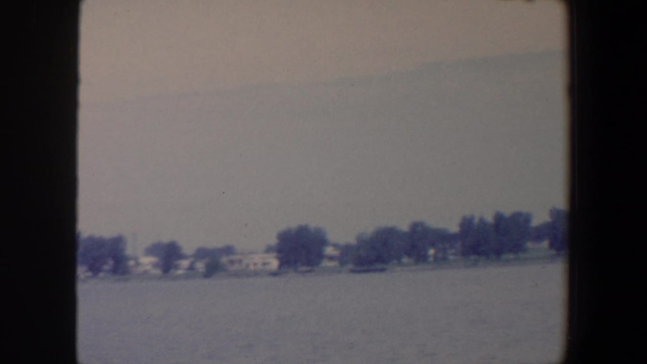 Distant tree-lined shoreline and small houses across calm lake water under a gray sky.