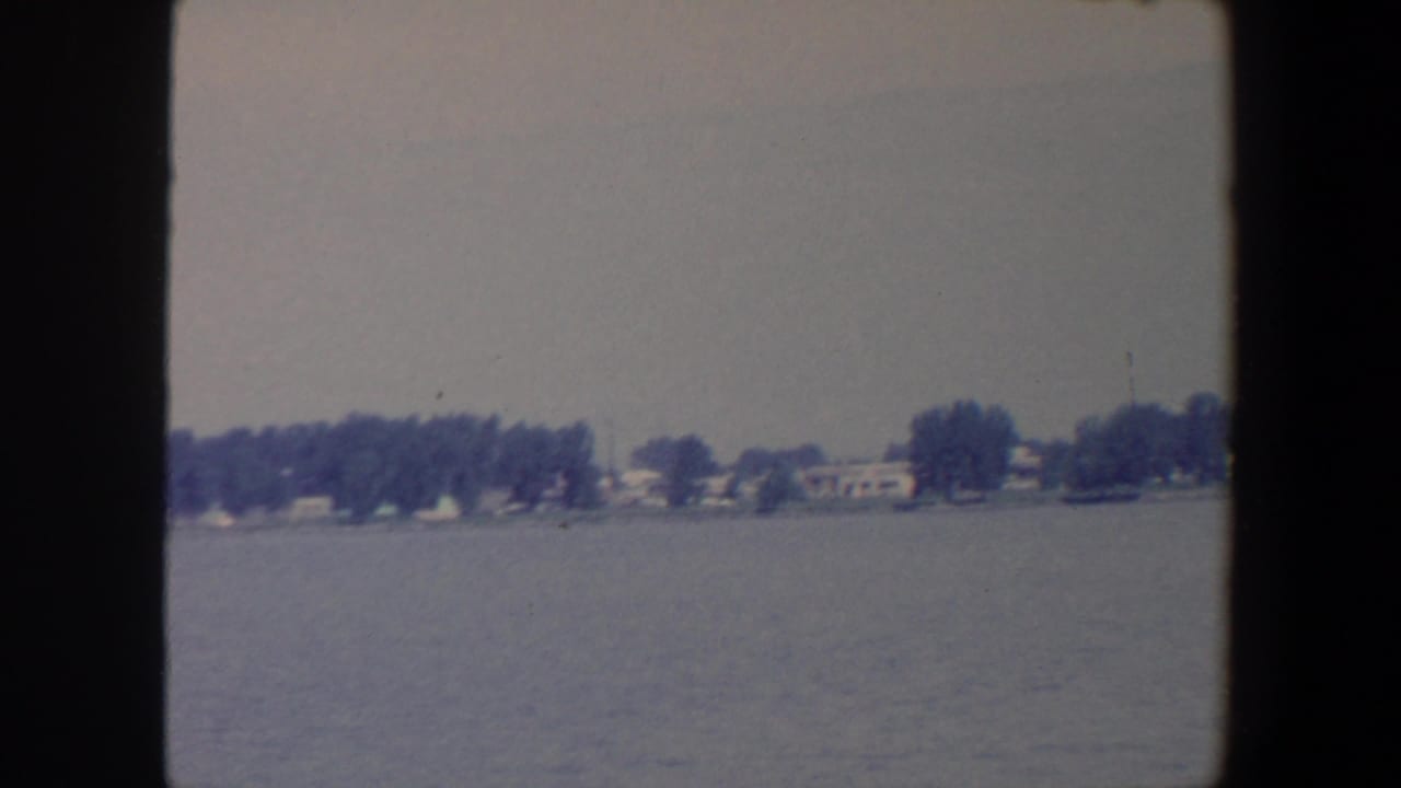 1959 Iowa home-movie view across a lake toward a low shore with dense trees and scattered houses.