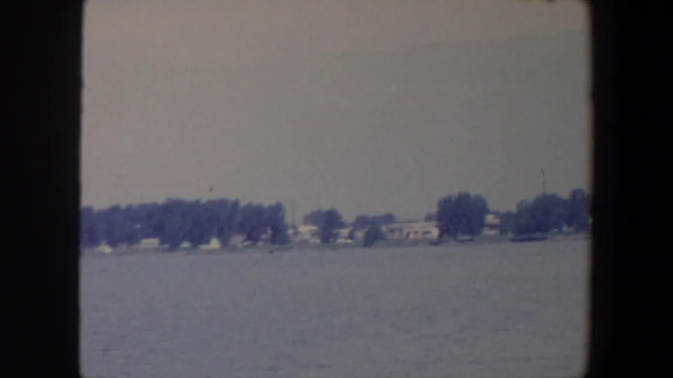 1959 Iowa home-movie view across a lake toward a low shore with dense trees and scattered houses.