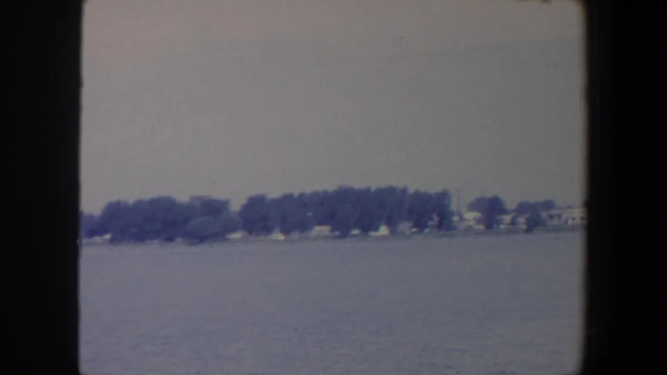1959 Iowa 8mm frame with muted lake water, a wooded shoreline, and pale houses partly screened by trees.
