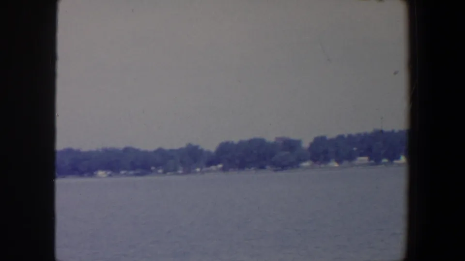 1959 Iowa lakeside frame showing calm water, a low tree-lined shore, and small distant houses under gray light.