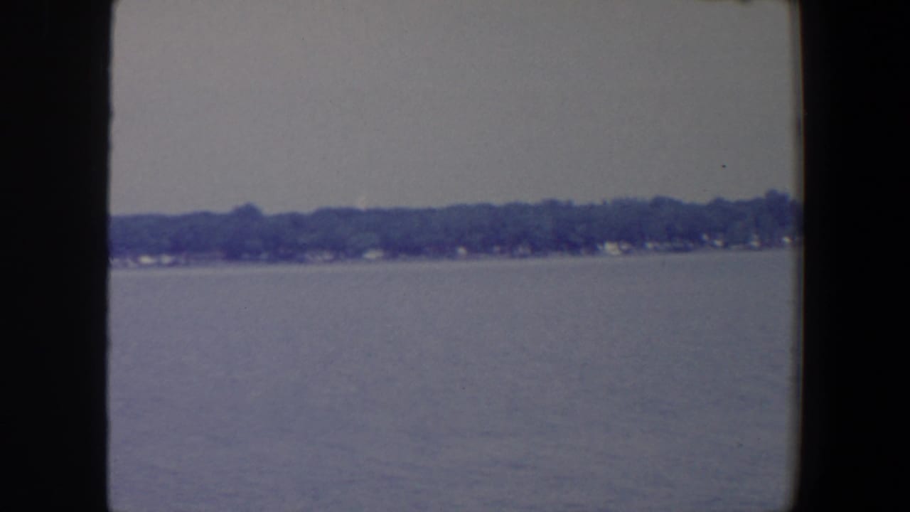 Boat-view frame of a broad Iowa lake or lagoon, with trees and small structures along the far shore.