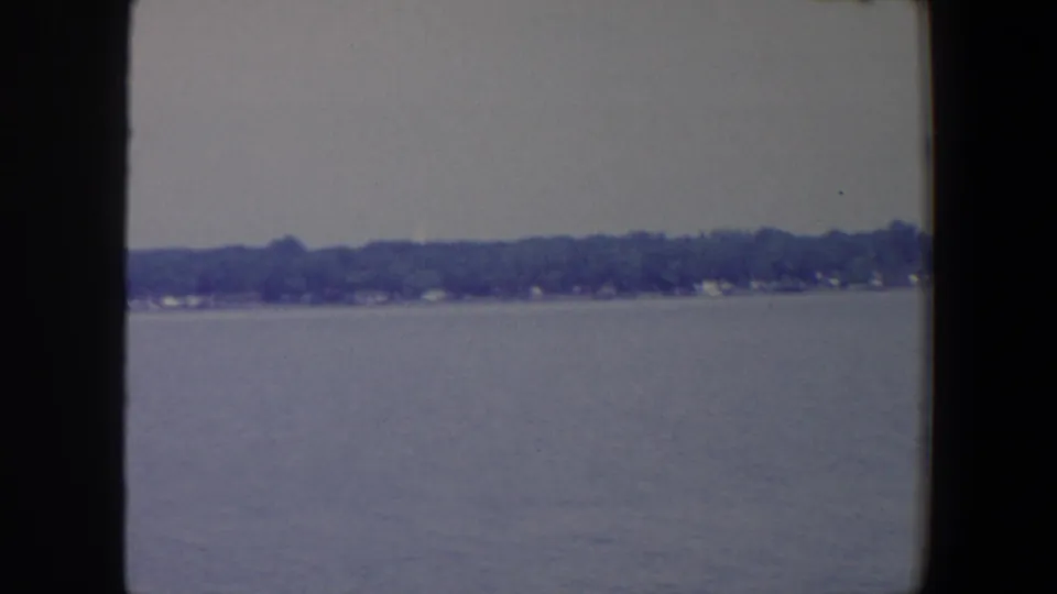 Boat-view frame of a broad Iowa lake or lagoon, with trees and small structures along the far shore.