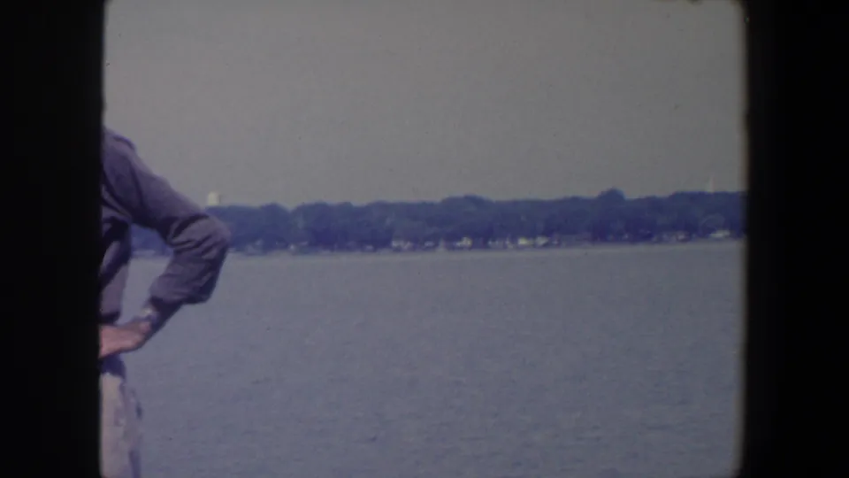 1959 Iowa boat-trip frame with a partial adult figure in a long-sleeved shirt against the lake and shoreline.
