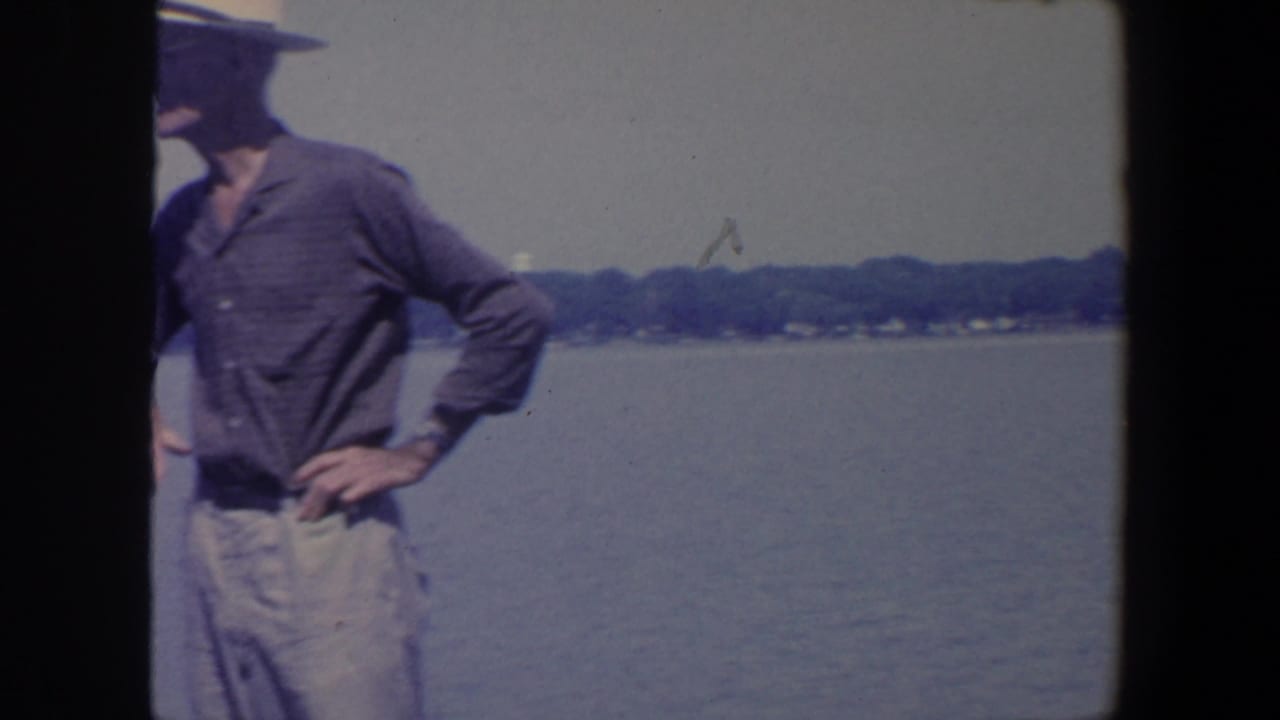 Man in a hat stands by calm water with a distant tree line behind him.