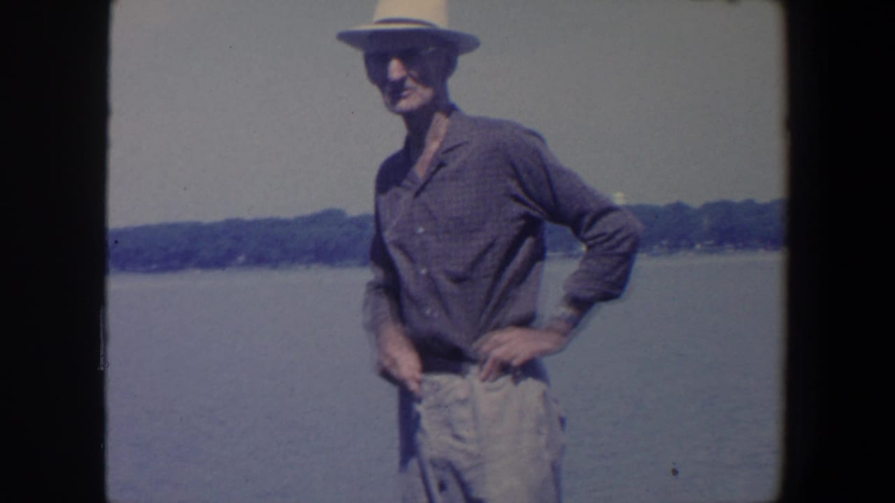 Man in a straw hat stands with one hand on his hip beside the lake.