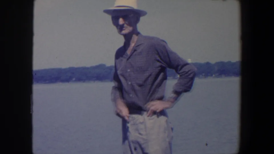 Adult man in a straw hat and casual shirt stands near water in Iowa home-movie footage from 1959.
