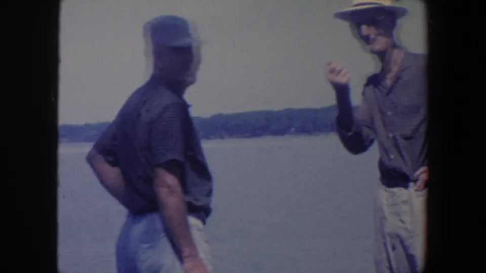 Two adult men stand near an Iowa lake, one facing away while the other gestures during an informal 1959 home-movie moment.