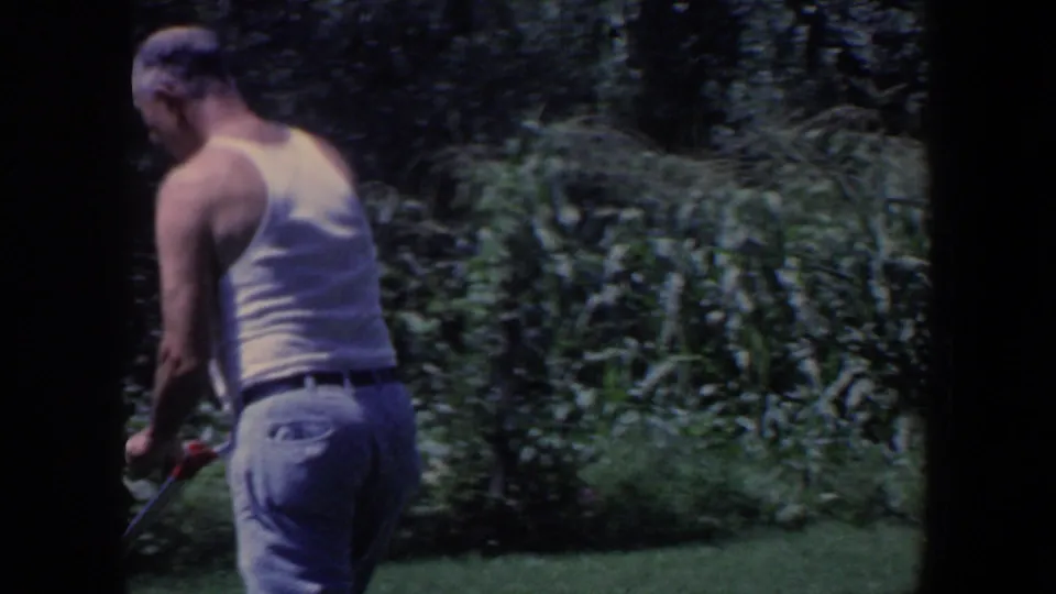 Adult man in a white undershirt and jeans moves along a lawn beside dense vegetation in the same Iowa home-movie clip.