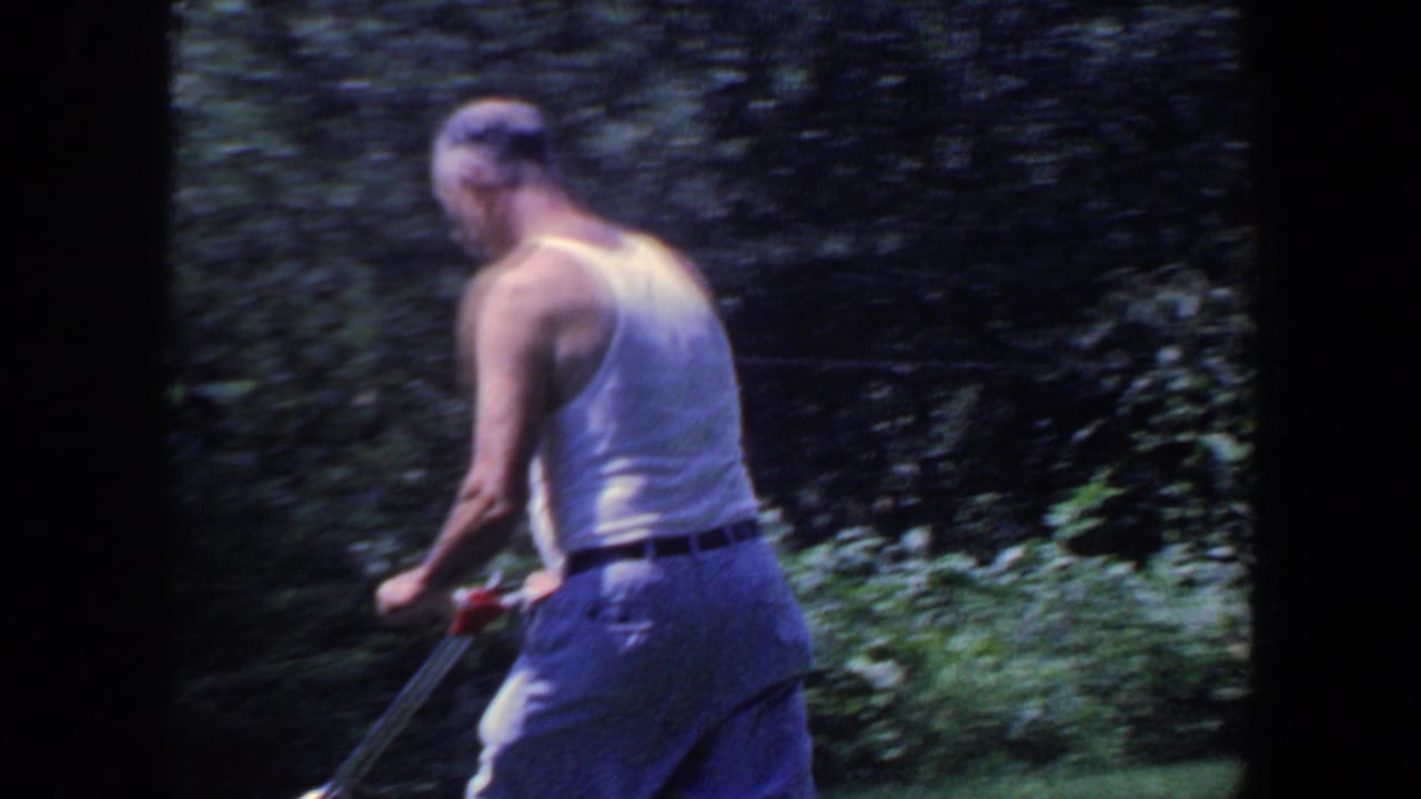 Man in a white tank top pushes a mower through a leafy yard.
