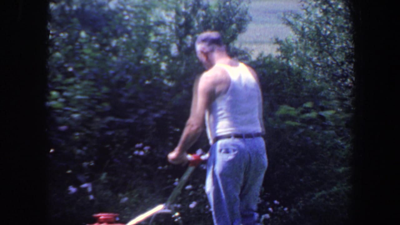 Man seen from behind guiding a mower beside bushes and trees.