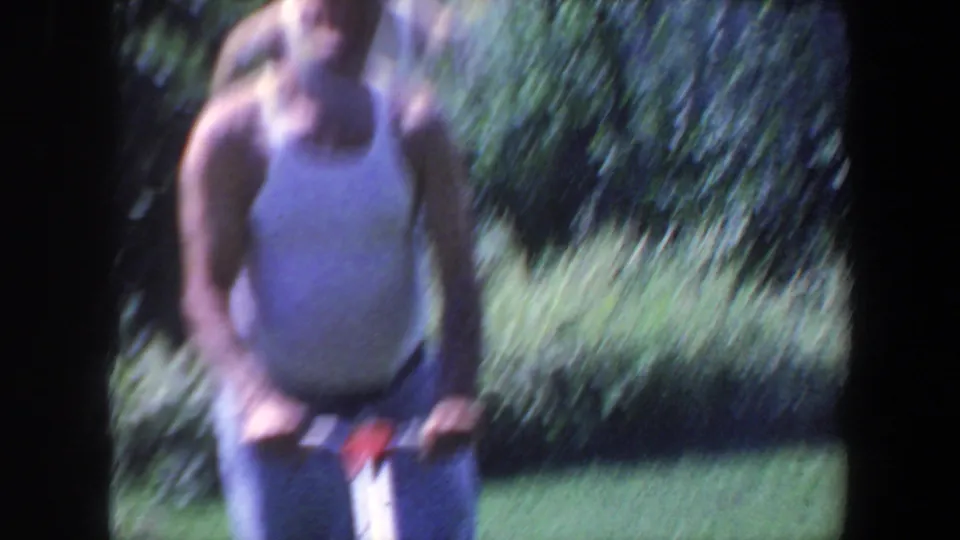 A softly focused 1959 Iowa frame shows the man facing the camera with mower handles in the foreground.