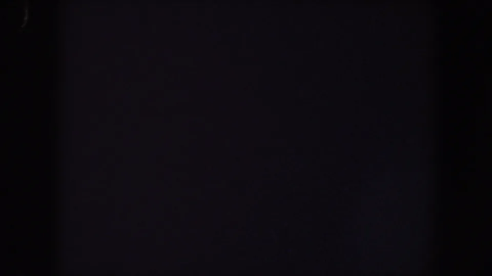 Near-black 1959 Iowa 8mm frame with faint blocky shadow shapes, visible grain, and a small pale speck near the lower left.