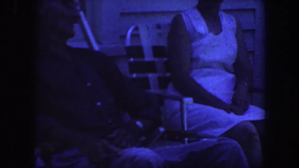 1959 Iowa home-movie frame showing two seated adults on folding chairs outside house siding, tinted blue by the film scan.