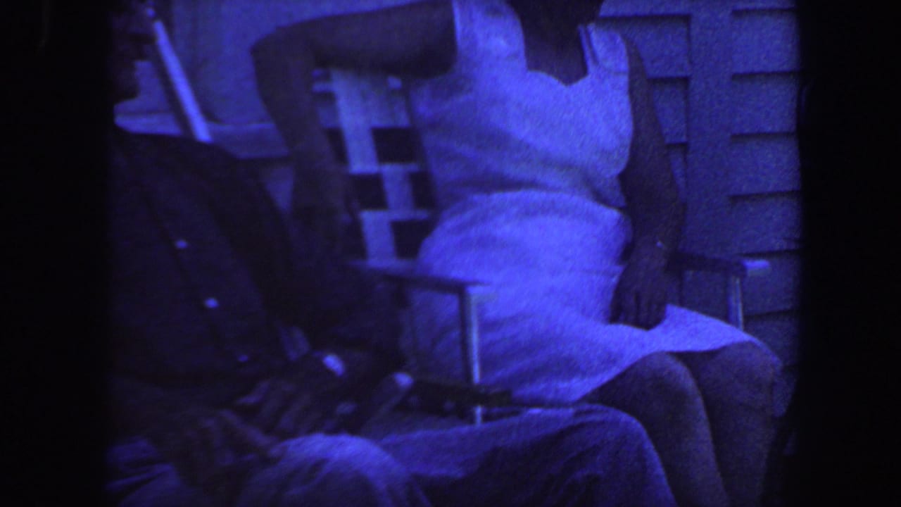 A woman in a white dress sits on a folding chair beside a partially visible adult.