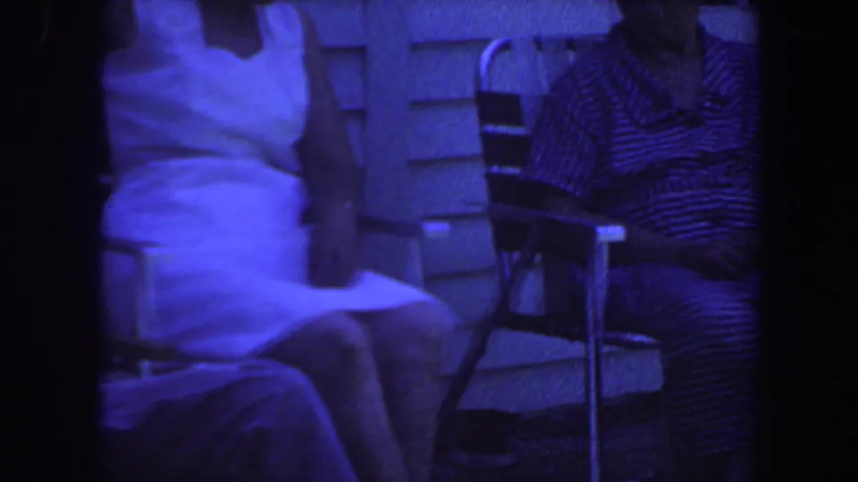 Blue-tinted 1959 Iowa home-movie frame showing seated adults outside a house, including a white dress and striped clothing.