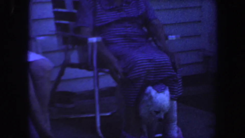 1959 Iowa home-movie frame of a seated person on a porch with a small white dog close to their knees and a house exterior behind.