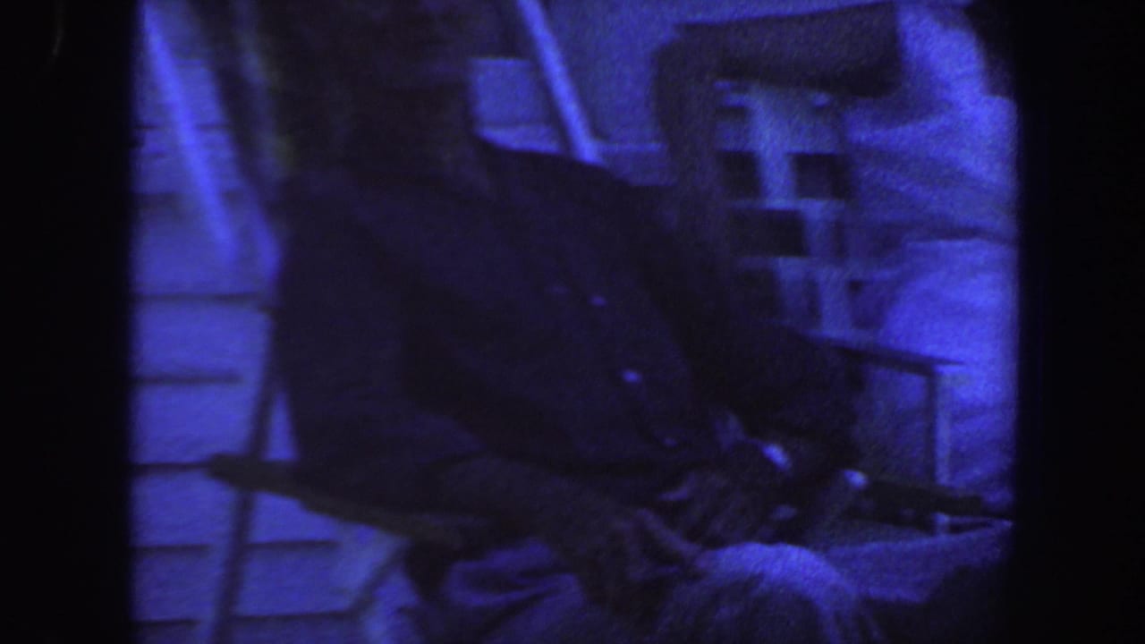 Seated adult in a lawn chair beside house siding in blue-tinted low light.