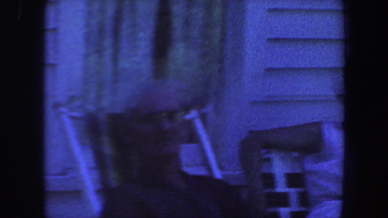 1959 Iowa home-movie frame showing lawn chairs near house siding, with the scene partly obscured by dark film borders.