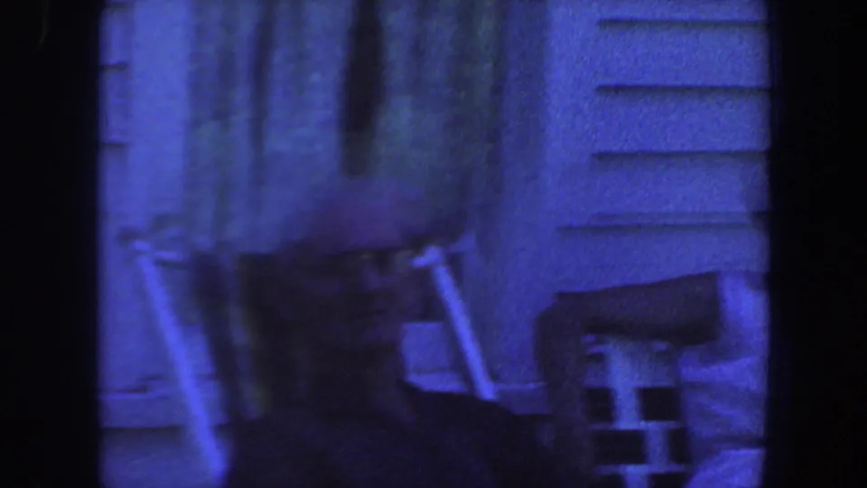 1959 Iowa home-movie frame showing lawn chairs near house siding, with the scene partly obscured by dark film borders.
