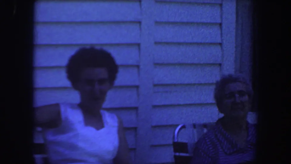 1959 Iowa 8mm frame showing two adults seated outdoors by house siding, one in a white sleeveless top.