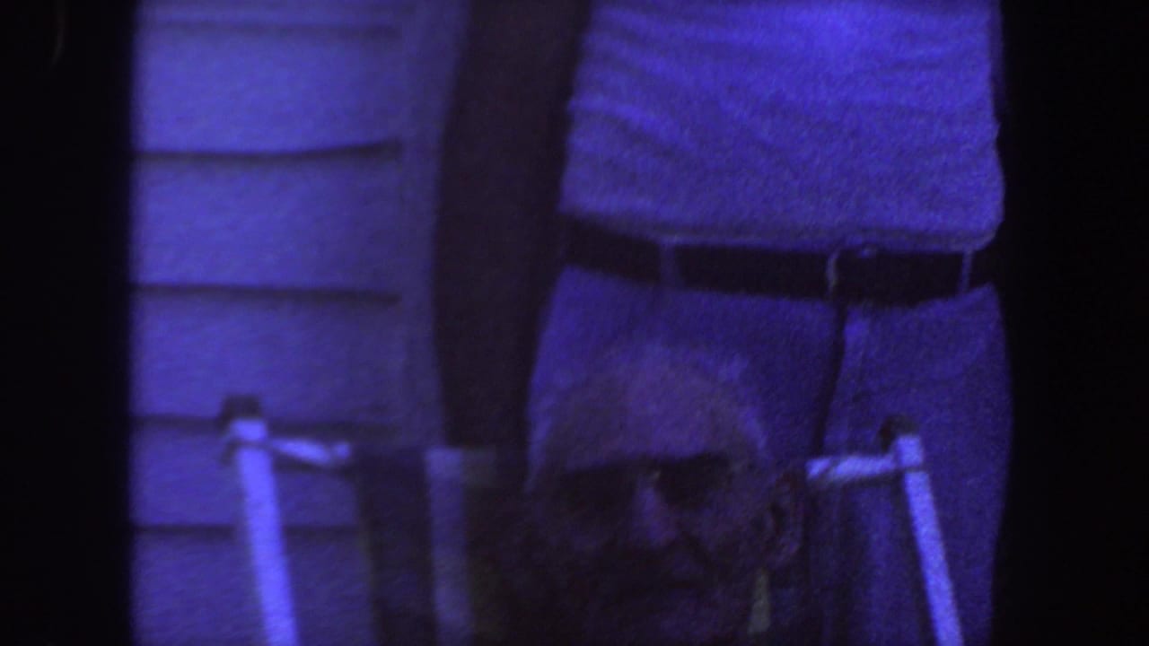 Man in a white tank top and belted trousers standing near house siding.